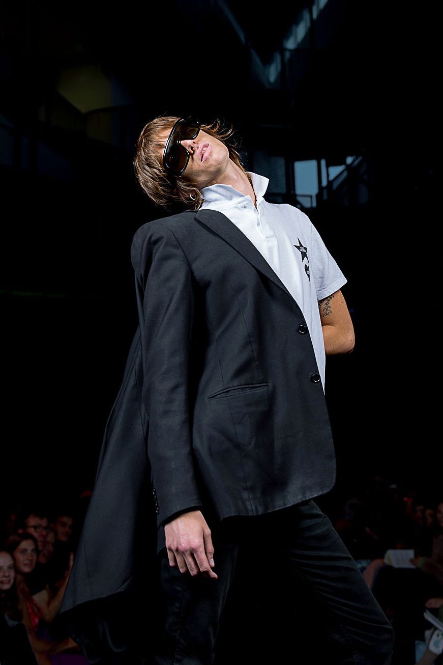Student modeling for the CC Fashion Show wearing a casual white tshirt with a black blazer hanging on one arm and dark sunglasses.