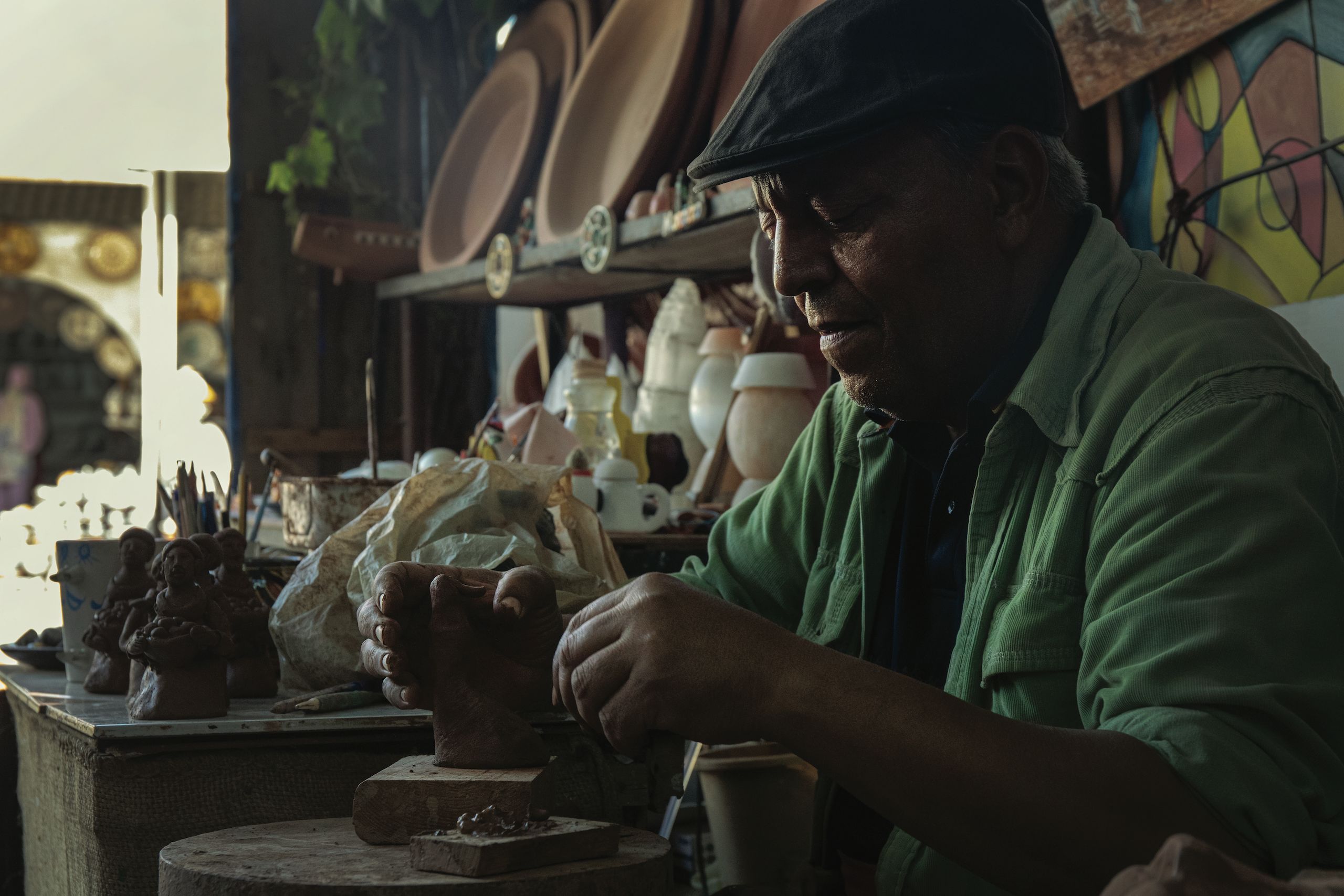 Man seated at his work station creating pottery with shelves of finished items behind him.